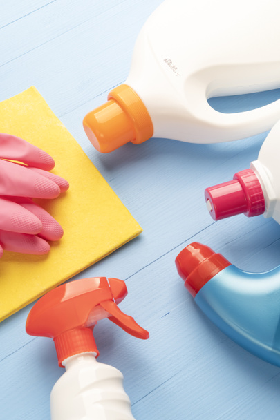 Для домашнего задания, моющие средства в ярких пластиковых бутылках и желтая тряпка с розовыми защитными перчатками крупным планом на деревянном синем фоне
. - Фото, изображение