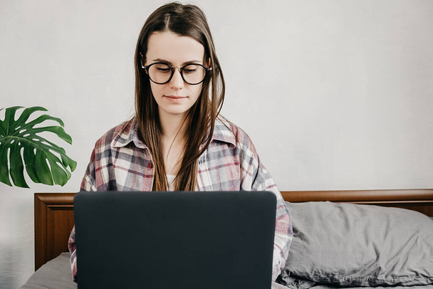Thoughtful woman in eyeglasses thinking about project, working on financial report, using laptop, sitting on bed, female employee pondering business strategy, solving problem. Social distancing - Fotoğraf, Görsel
