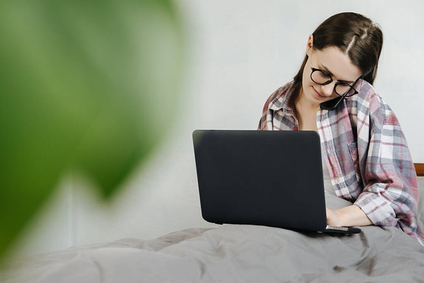 Smiling young woman wearing glasses talking on phone, sitting in bed at home with laptop, friendly manager consulting customer by smartphone, happy girl chatting with friends distracted from work - Photo, Image