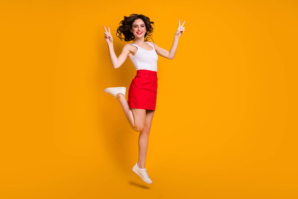 Full body profil fotografie legrační dáma skok vysoká dobrá nálada slunečný den víkend ukazuje V-znamení gesto oblečení bílá ležérní singlet červená krátká sukně boty izolované žlutá barva pozadí - Fotografie, Obrázek