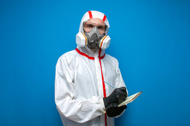 trabajador de desinfección en traje protector y respirador escribe estadísticas, médico virólogo en un fondo aislado azul
 - Foto, Imagen