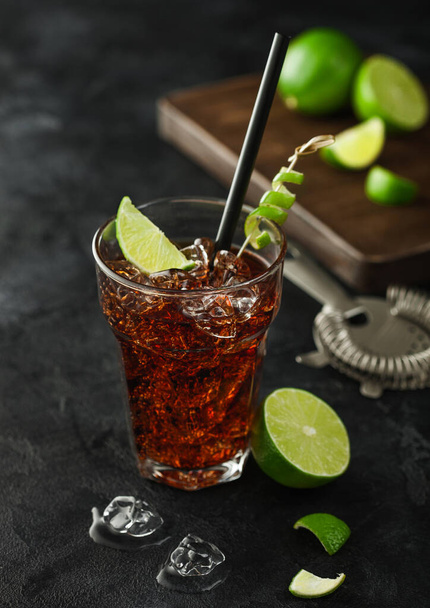 Cuba Libre cocktail in heavy crystal glass with ice and lime slice and peel, with straw and fresh limes with bar strainer on black table background. - 写真・画像