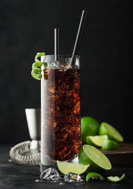 Cuba Libre cocktail in highball glass with ice and lime peel with straw and fresh limes and strainer on black table background. - 写真・画像