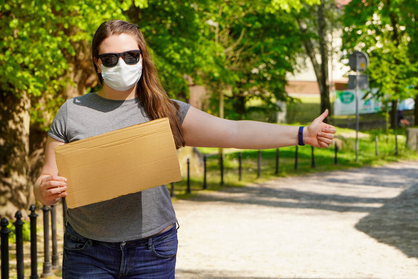 Girl in medical mask with empty cardboard sign stands by the road, catching car for Humorous hitchhiking trip. Getting out of quarantine, traveling after coronavirus. Pandemic fear. - Photo, Image