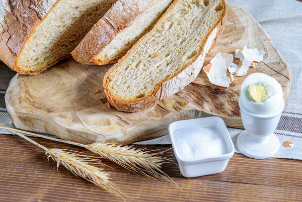 Pão integral fatiado, espiguetas de trigo, sal e ovo cozido na mesa de madeira da cozinha. Alimentos saudáveis para comer no campo
.  - Foto, Imagem