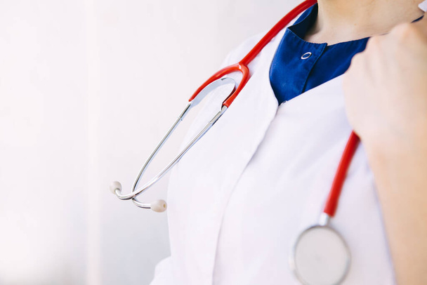 Красный стетоскоп на шее молодой женщины-врача в белом халате и хирургическом костюме перед пациентом с болезнью легких, вирусом, коронавирусом, копировальным пространством, рыжими волосами, синим костюмом, униформой
 - Фото, изображение