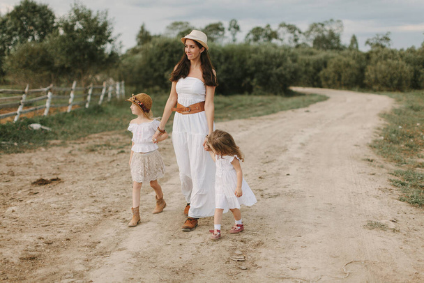 Mom with 2 daughters walks the village in white dresses on a summer day, life on the farm - Фото, изображение