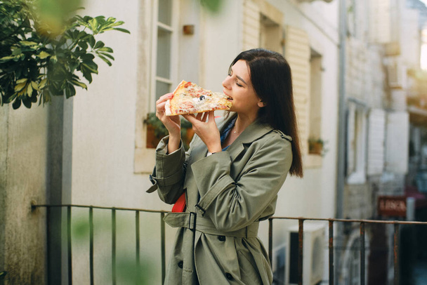 Beautiful brunette woman in a green coat eats pizza and walks around the city - Fotoğraf, Görsel