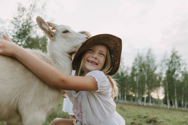 children and goat in the meadow on a summer day, feeding the animal, life on the farm - Zdjęcie, obraz