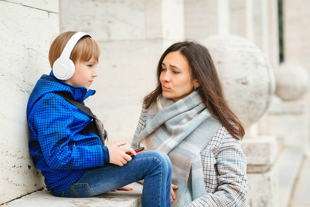 Smutny chłopak słucha muzyki na spacerze. Matka i syn spędzają razem czas na zewnątrz. Znudzony chłopak używający bezprzewodowych słuchawek. Macierzyństwo, związek i rodzina. Nowoczesna technologia, styl życia. - Zdjęcie, obraz
