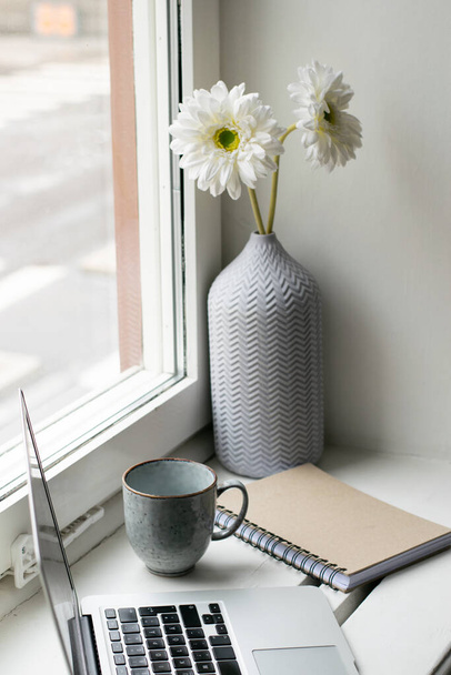 Heldere werkplek bij het raam met bloemen, laptop, notebook en koffiebeker - Foto, afbeelding
