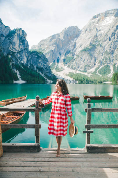 woman in red sundress sitting on wooden stairs looking at lake in mountains. travel concept - Photo, Image