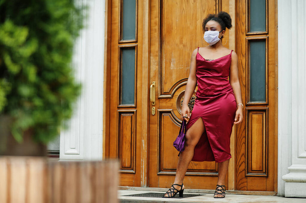 Covid-19, besmettelijk virus. Afrikaanse vrouw met krullend haar, draagt rode zijden jurk en medisch wegwerpmasker, geeft om haar gezondheid en beschermt in een gevaarlijke situatie. - Foto, afbeelding