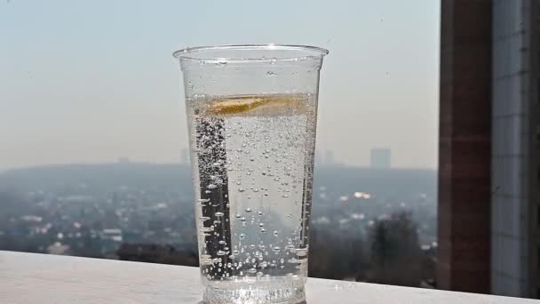 video a slice of lemon falls into a transparent plastic glass with soda. A refreshing cocktail by the window overlooking the city. Hot summer day. - Séquence, vidéo