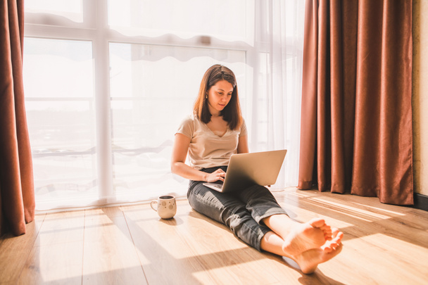 femme assise sur le sol travaillant sur un ordinateur portable près d'une grande fenêtre. isolement personnel. quarantaine. télétravail
 - Photo, image