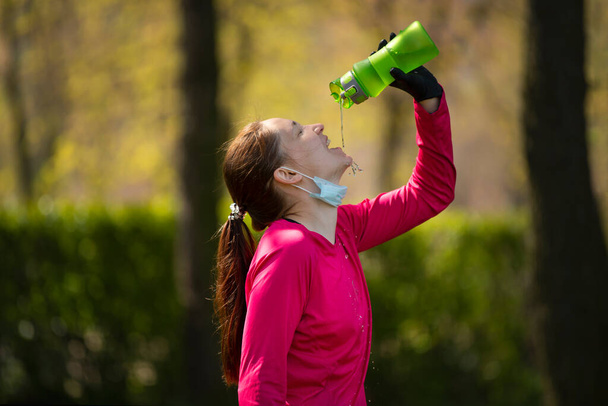 giovane ragazza beve acqua dopo aver praticato sport nel parco. Ragazza in maschera medica e guanti dopo la quarantena
 - Foto, immagini