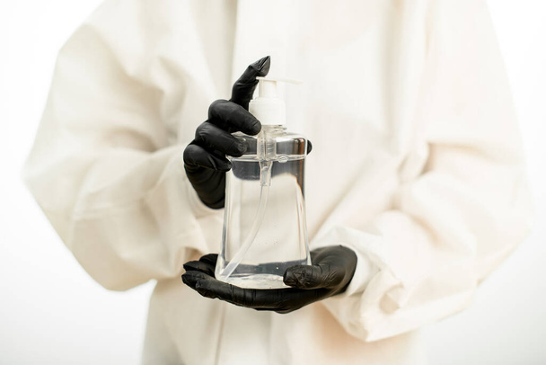 Close-up of transparent bottle with disinfectant in hands of person in black gloves and white protective suit - Valokuva, kuva