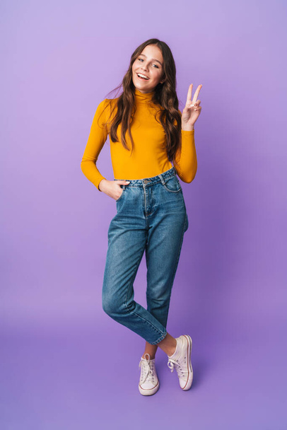 Full length image of young beautiful woman with long brown hair smiling and gesturing peace fingers isolated over violet background - Valokuva, kuva