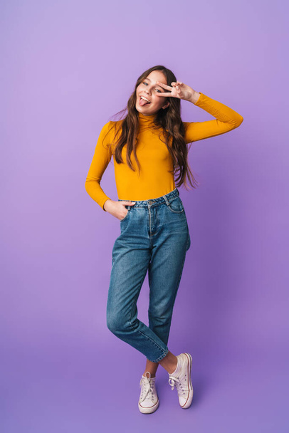 Full length image of young beautiful woman with long brown hair smiling and gesturing peace fingers isolated over violet background - Foto, Imagem