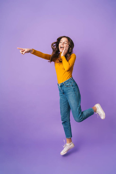 Full length image of young beautiful woman with long brown hair smiling and pointing finger at copyspace isolated over violet background - Foto, afbeelding