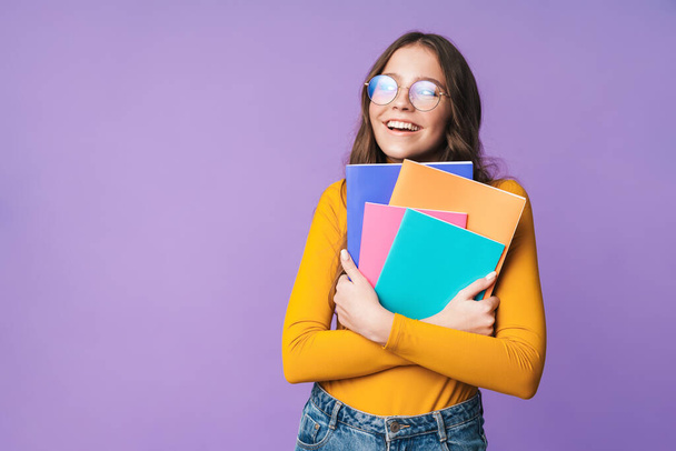 Imagen de una joven estudiante hermosa con anteojos sonriendo y sosteniendo libros de ejercicios aislados sobre fondo violeta
 - Foto, Imagen