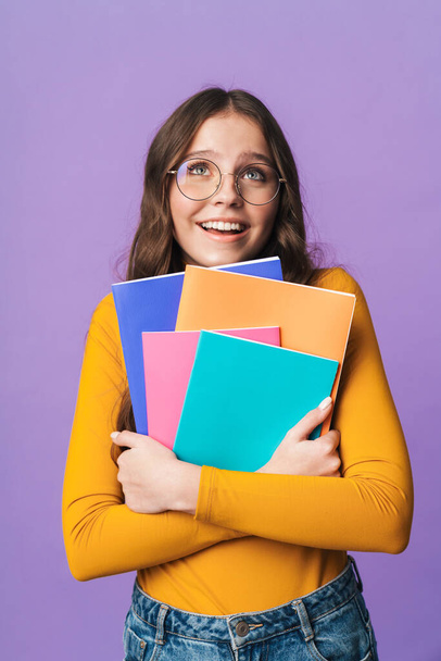 Image of young beautiful student girl wearing eyeglasses smiling and holding exercise books isolated over violet background - Fotoğraf, Görsel
