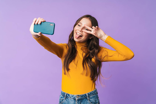 Image of young beautiful woman with long brown hair smiling and taking selfie photo on cellphone isolated over violet background - Фото, зображення