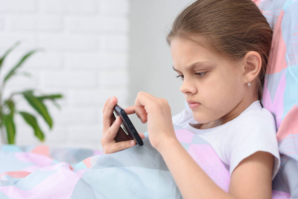 女の子はベッドに横たわっている間、携帯電話で遊んで - 写真・画像