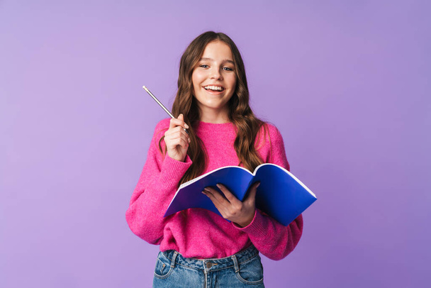 Image of young beautiful student girl smiling and holding exercise book isolated over violet background - Zdjęcie, obraz