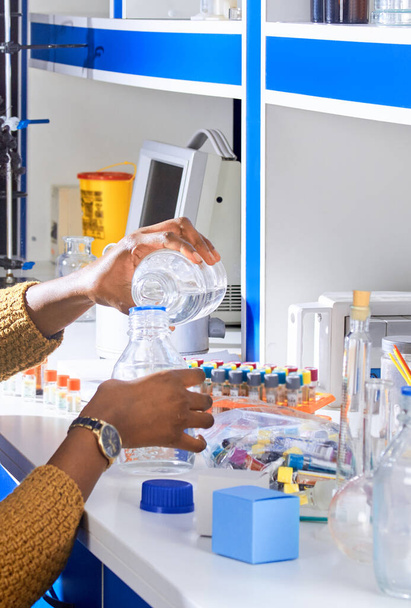 Nuori afrikkalainen naispuolinen tech tai tiedemies kaataa puskuriliuosta pullosta pulloon verikokeiden määritykseen nykyaikaisessa laboratoriossa tai tutkimuslaitoksessa. - Valokuva, kuva