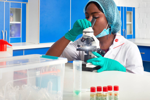 Afrikai női orvostanhallgató, női tudós Védő fehér köpenyben, műanyag kalapban, kesztyűben, maszkban mikroszkóppal működik laboratóriumban, tesztlaborban. A betegek biopsziás mintáinak elemzése. Anatómiai munka - Fotó, kép
