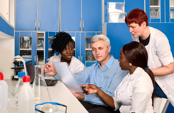 Kaukasische Senior-Professoren, junge afrikanische Studenten diskutieren die Ergebnisse im Labor. Multikulturelles Team aus Wissenschaftlern und Medizinern optimiert Schnelltest für neuartiges Coronavirus SARS-COV-2 im Krankenhaus. - Foto, Bild