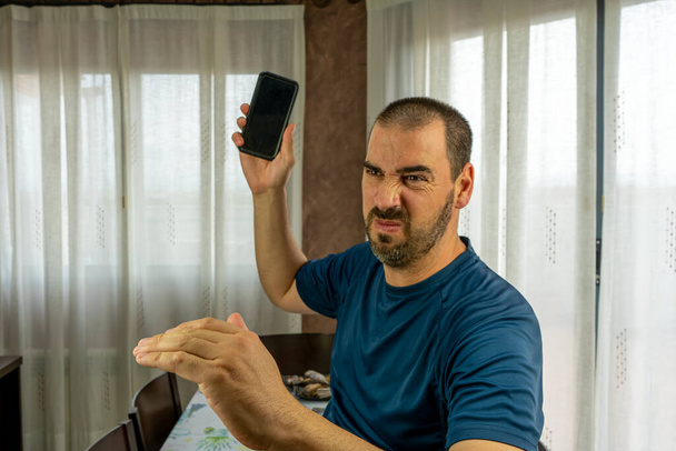 Разъяренный мужчина с бородой и короткими волосами в синей рубашке, бросающий мобильник в гостиную дома
 - Фото, изображение