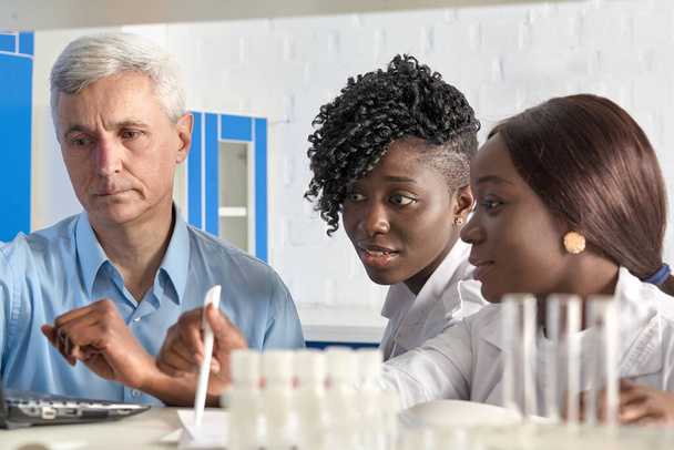 Europese teamleider en Afrikaanse geneeskundestudenten werken in testlaboratorium of onderzoeksfaciliteit en bespreken optimalisatie van nieuwe coronavirus testmethoden. Senior Kaukasische man en twee vrouwen. - Foto, afbeelding