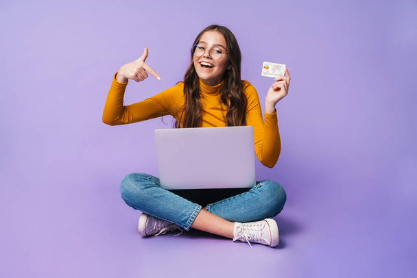 Image of young beautiful woman holding credit card and using laptop while sitting with legs crossed isolated over violet background - Zdjęcie, obraz