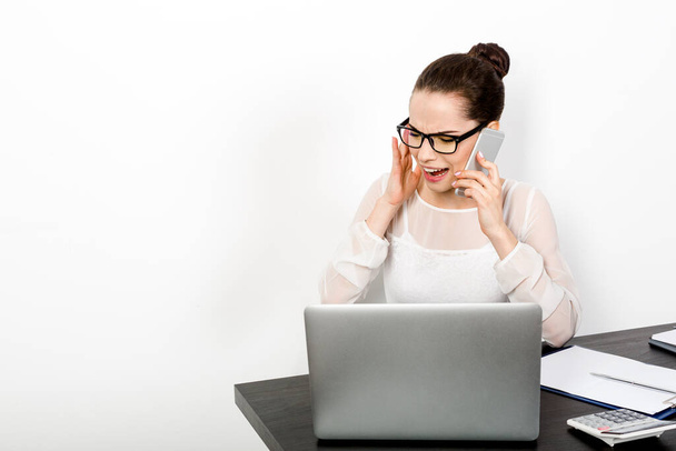 A young enerpreneur woman sits at her workplace with a laptop near a white wall while talking emotionally and loudly on the phone. The concept of successful business negotiations - Photo, Image