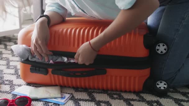 Młoda dziewczyna stojąca na łokciu na przepełnionej pomarańczowej walizce, próbująca ją zamknąć - Materiał filmowy, wideo