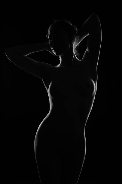 A naked girl stands in the pose of Venus. Photo of a gorgeous Nude female body in a contoured light on a dark background - Foto, afbeelding