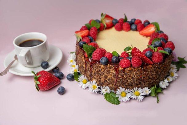Cheesecake versierd met bessen en bloemen op een licht roze achtergrond met een kopje koffie aan de linkerkant. - Foto, afbeelding