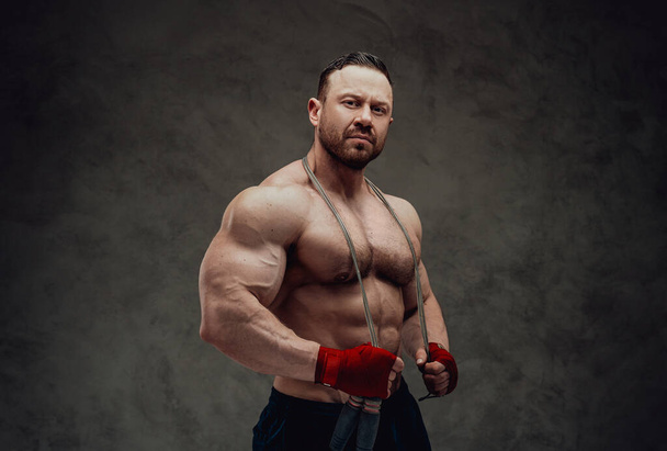 Симпатичный спортсмен с огромными мускулами в спортивной одежде позирует с скакалкой на шее и смотрит в камеру в темной студии
 - Фото, изображение