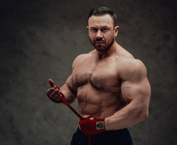 Seriöser und energiegeladener Fitnesstrainer, der konzentriert seine Muskeln in einem dunklen Studio zeigt, in Fitness-Shorts und Boxhandtaschen - Foto, Bild