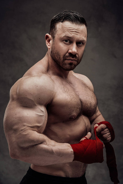 Hemdloser erwachsener männlicher Bodybuilder, der seinen großen Bizeps in einem dunklen Studio vor grauem Hintergrund zeigt und stark aussieht, während er seine Hände mit rotem Verband tapeziert - Foto, Bild