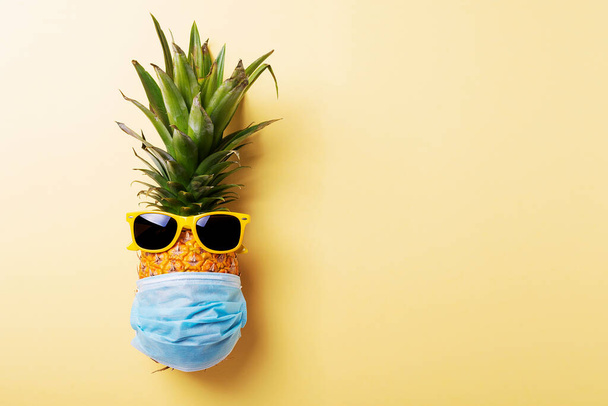 Concept of summer vacation 2020. Pine apple with a medical mask and sun glasses on the yellow background. Top view image - 写真・画像