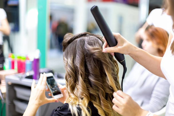 στυλίστρια close up κάνει ένα κορίτσι πελάτη styling μαλλιά κουνώντας με σίδερα κέρλινγκ σε ένα κομμωτήριο. πελάτης κορίτσι ψάχνει σε smartphone σε θολούρα. Κοντινό πλάνο, απαλή εστίαση - Φωτογραφία, εικόνα
