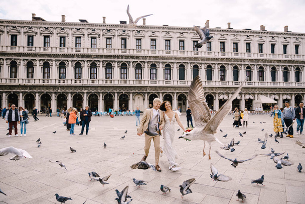 Ślub w Wenecji, Włochy. Panna młoda i pan młody biegną przez stado latających gołębi na Piazza San Marco, pośród Narodowego Muzeum Archeologicznego w Wenecji, otoczeni tłumem turystów. - Zdjęcie, obraz