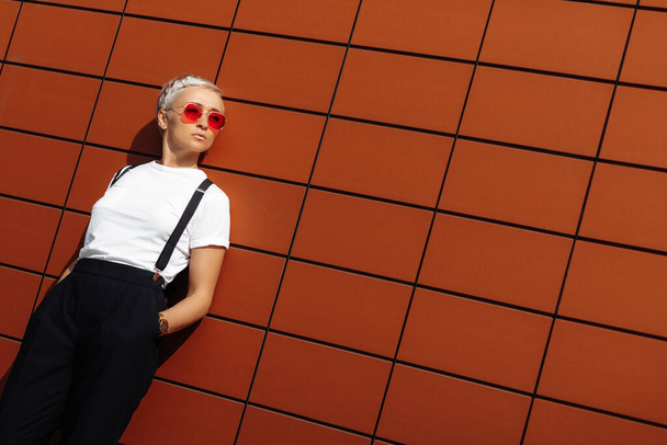 Beautiful hipster young woman wearing red sunglasses stay near wall of modern building. - Photo, image