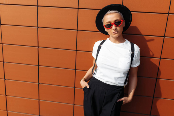 Serious hipster young woman wearing red sunglasses and black hat stay near wall of modern building. - 写真・画像