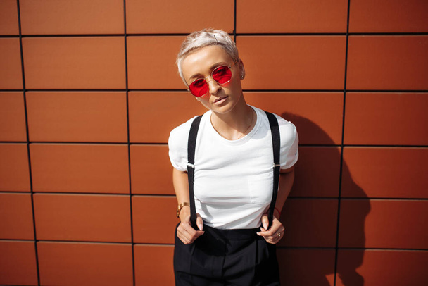 Stylish hipster young woman wearing red sunglasses stay near wall of modern building. - Фото, изображение