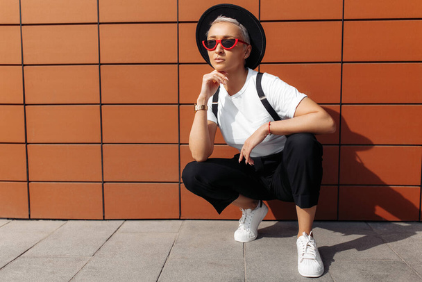 Hipster young woman wearing red sunglasses and black hat sitting near wall of modern building. - Фото, зображення