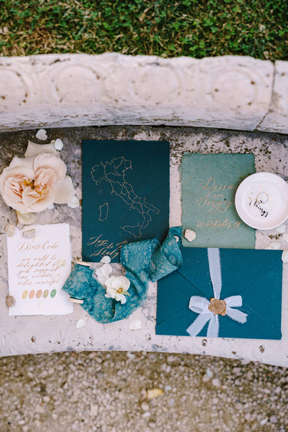 Blaue Attrappe von Hochzeitseinladungen, blauer Umschlag mit Siegeldruck aus Siegelwachs, ein seidenblaues Band, ein weißes Laken mit den Farben des Dessous-Codes, rosa Blume vor Marmorsteinhintergrund - Foto, Bild